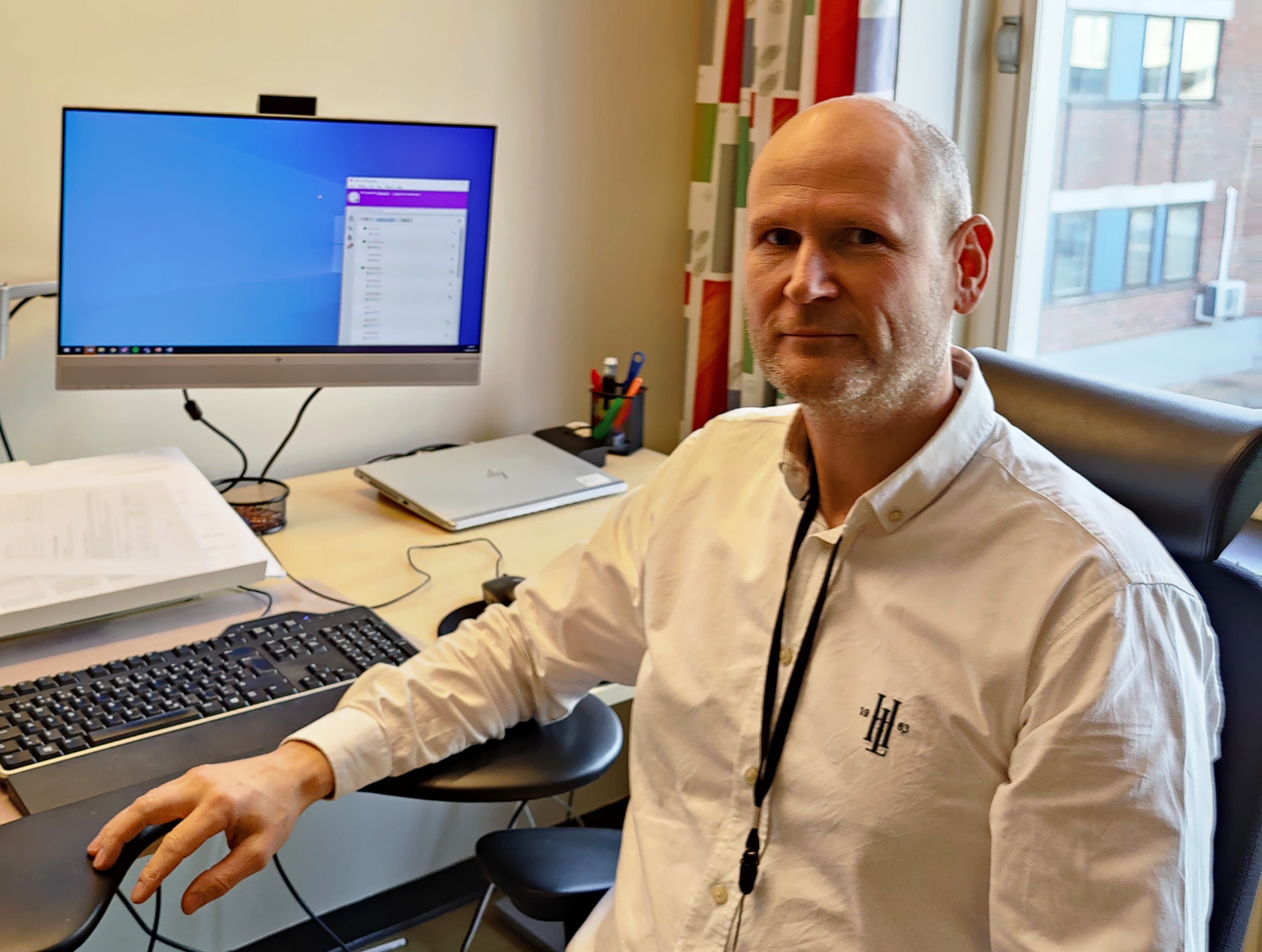 Person med vit skjorta sitter vid ett skrivbord, personen kollar in i kameran.